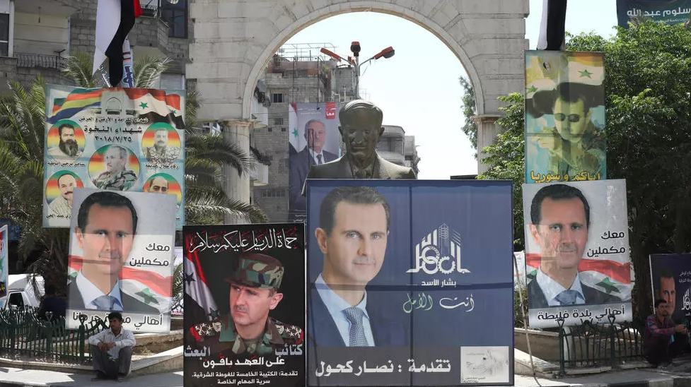 رجل يجلس بجوار تمثال للرئيس السوري السابق حافظ الأسد محاطا بملصقات انتخابية لنجله الرئيس السوري بشار الأسد في دمشق